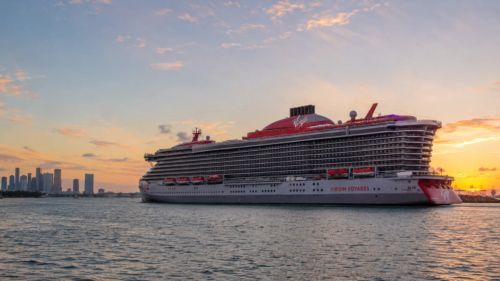 Virgin Voyages cruise ship passing a city skyline at sunset