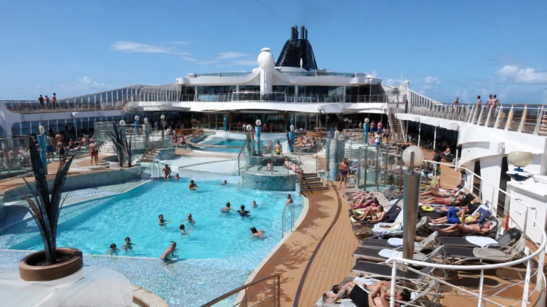 Open cruise ship pool deck with swimmers and rows of loungers