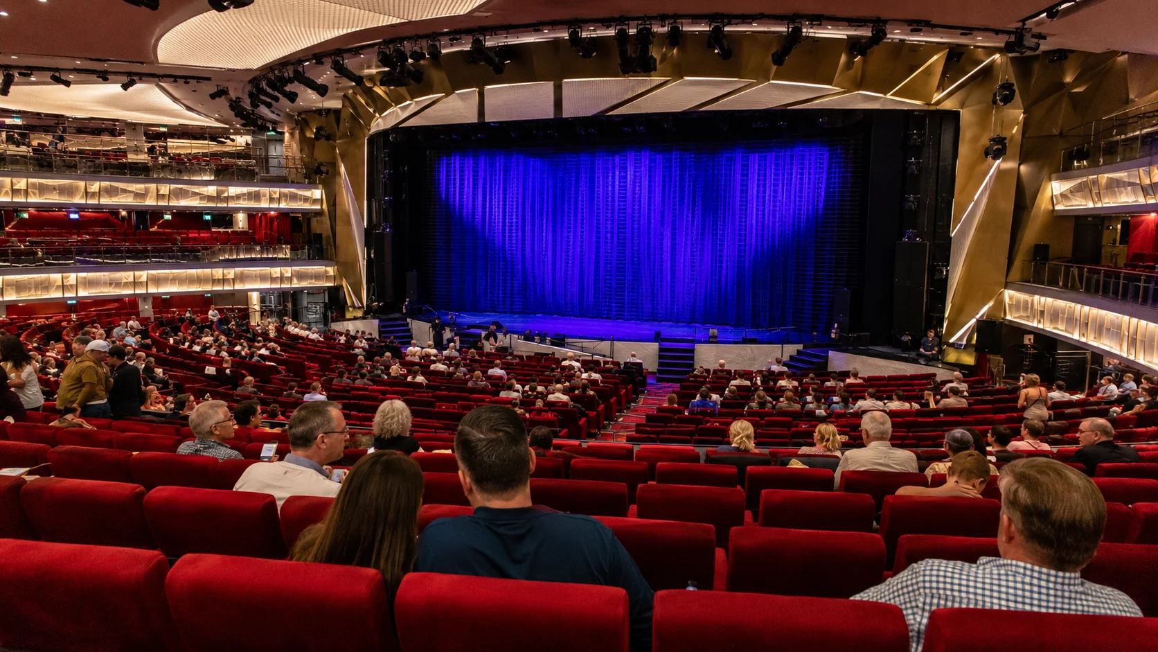 Audience seated in a red cruise ship theater before a purple-lit stage