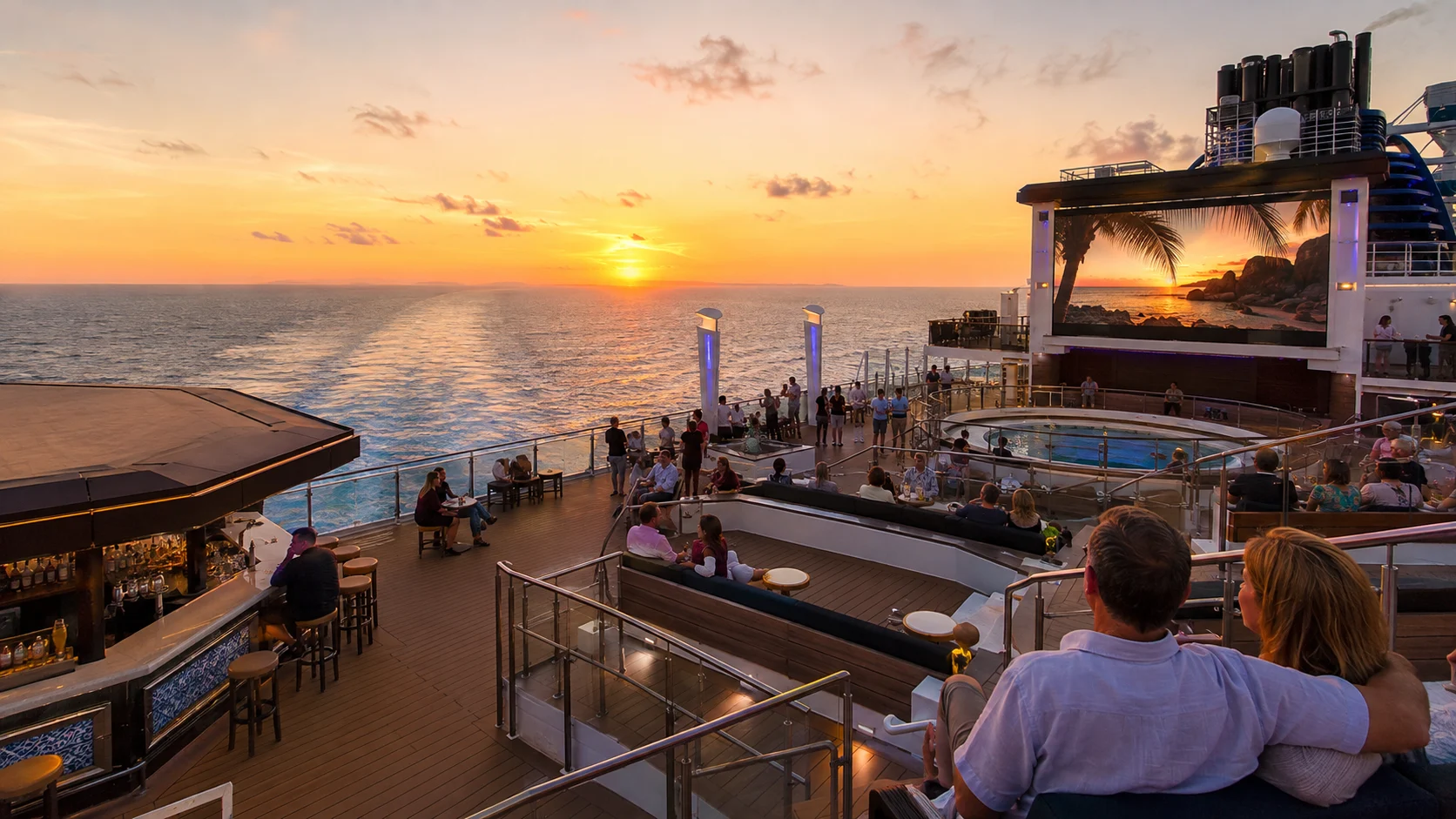Crowded outdoor cruise deck overlooking a stage and movie screen at sunset