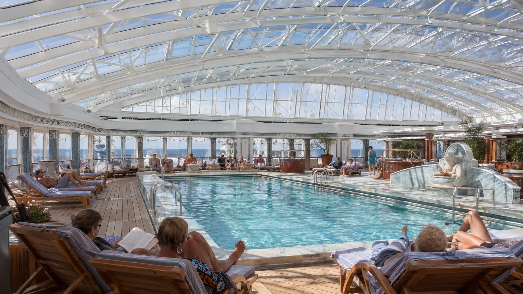 Passengers relaxing beside a glass-roofed indoor cruise ship pool