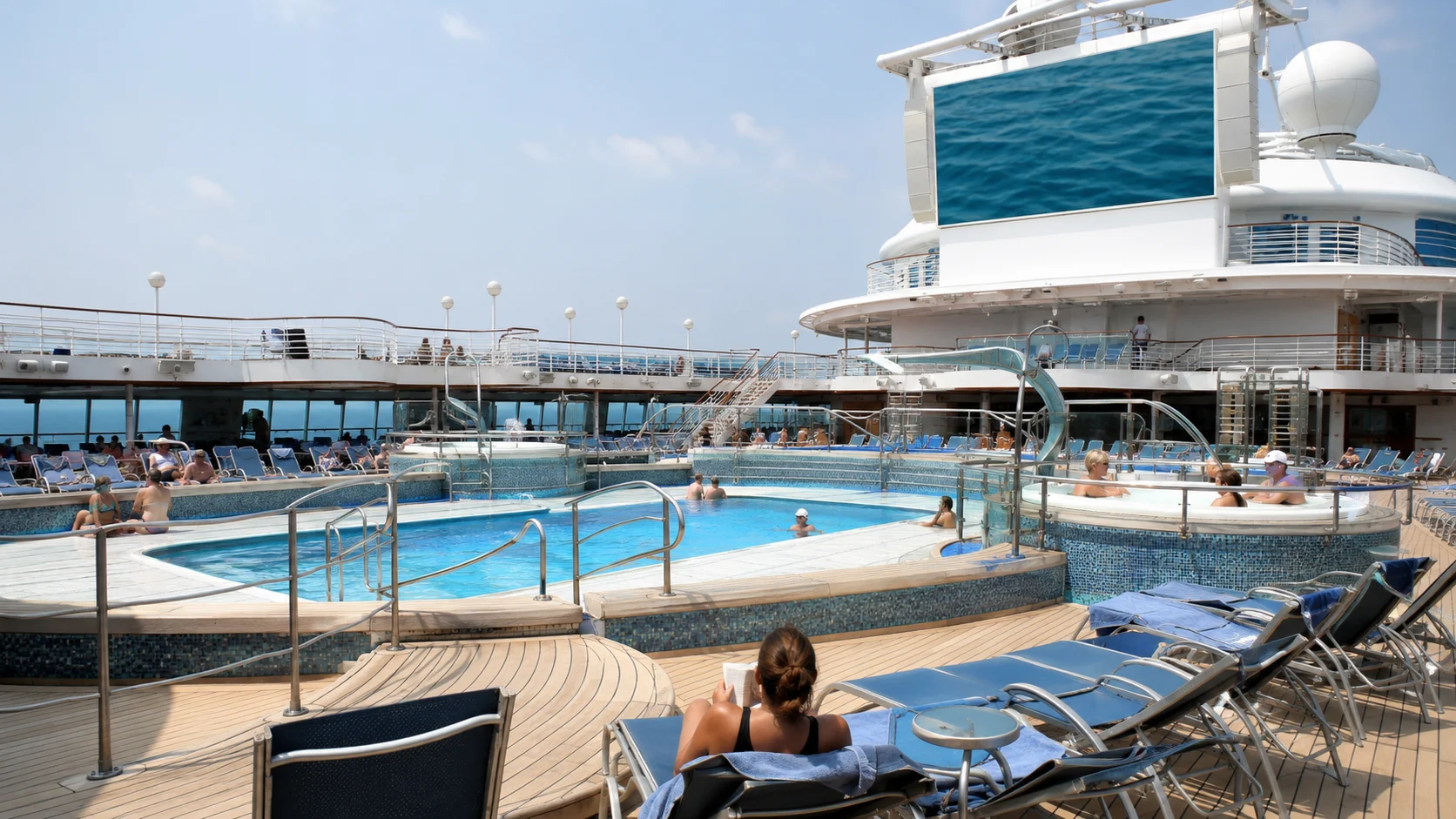 Cruise ship pool deck with blue loungers and a large outdoor screen