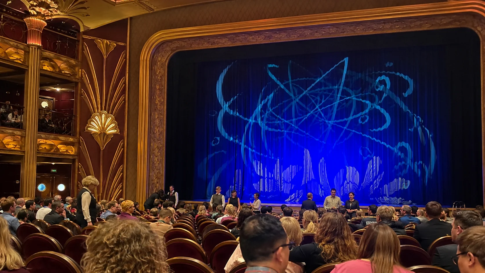 Audience watching a blue-lit performance in the Walt Disney Theatre aboard Disney Dream