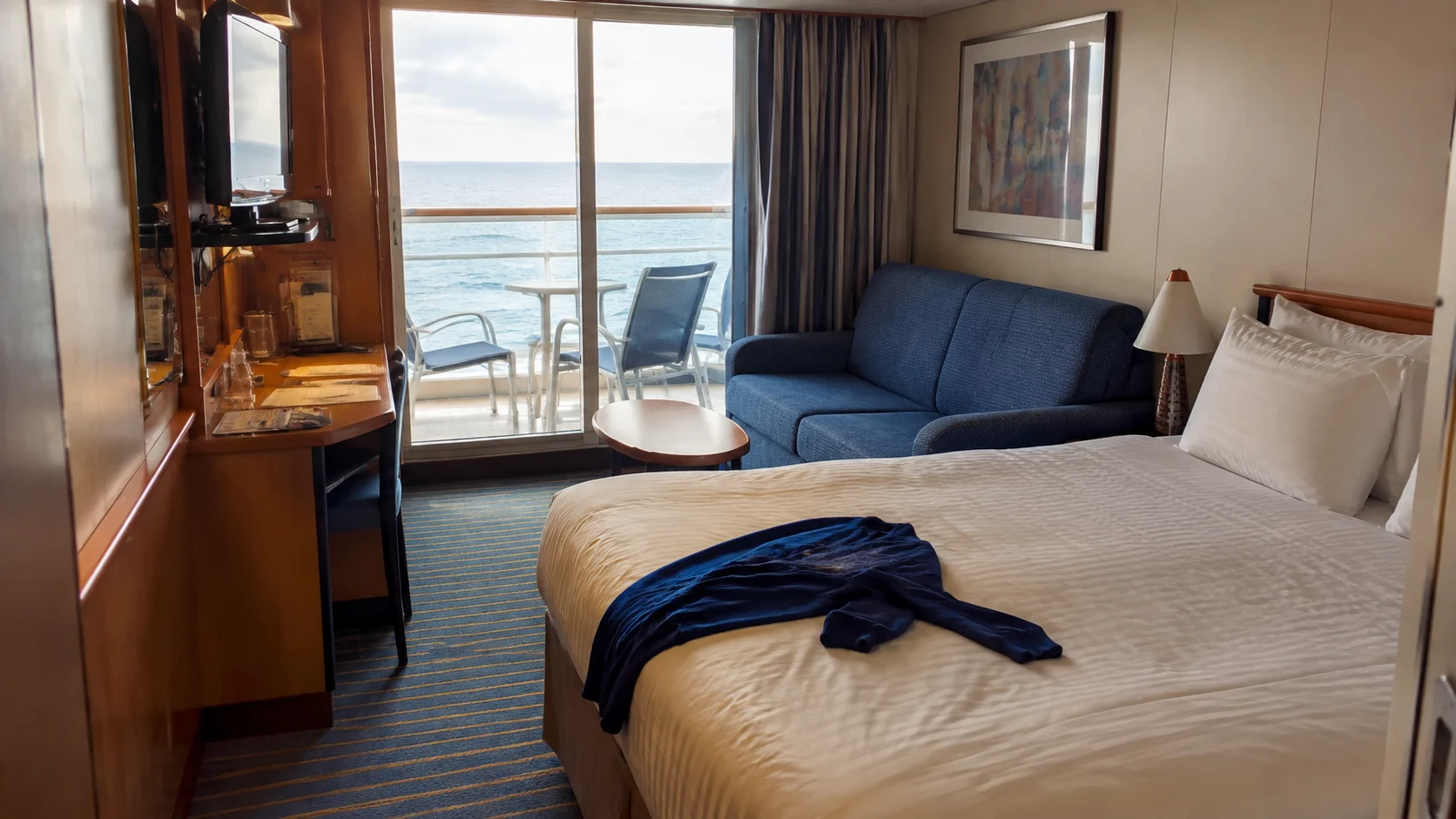 Cruise cabin with a double bed, blue sofa, and ocean-view balcony