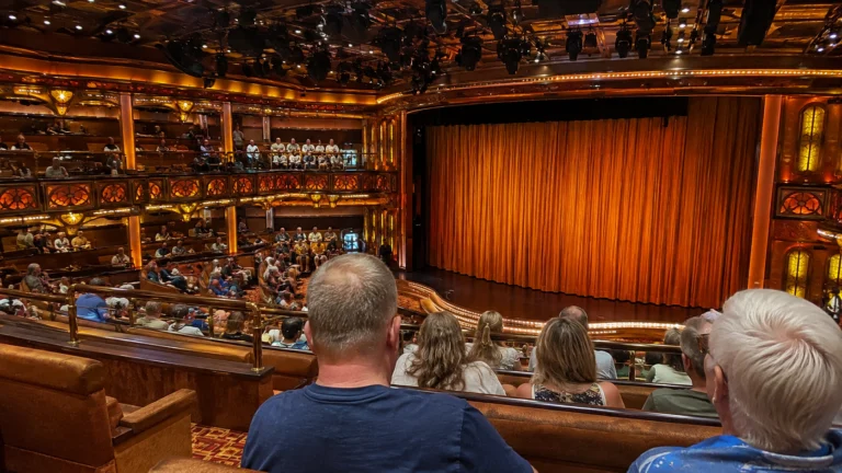 Audience seated before an orange stage curtain in an ornate cruise ship theater