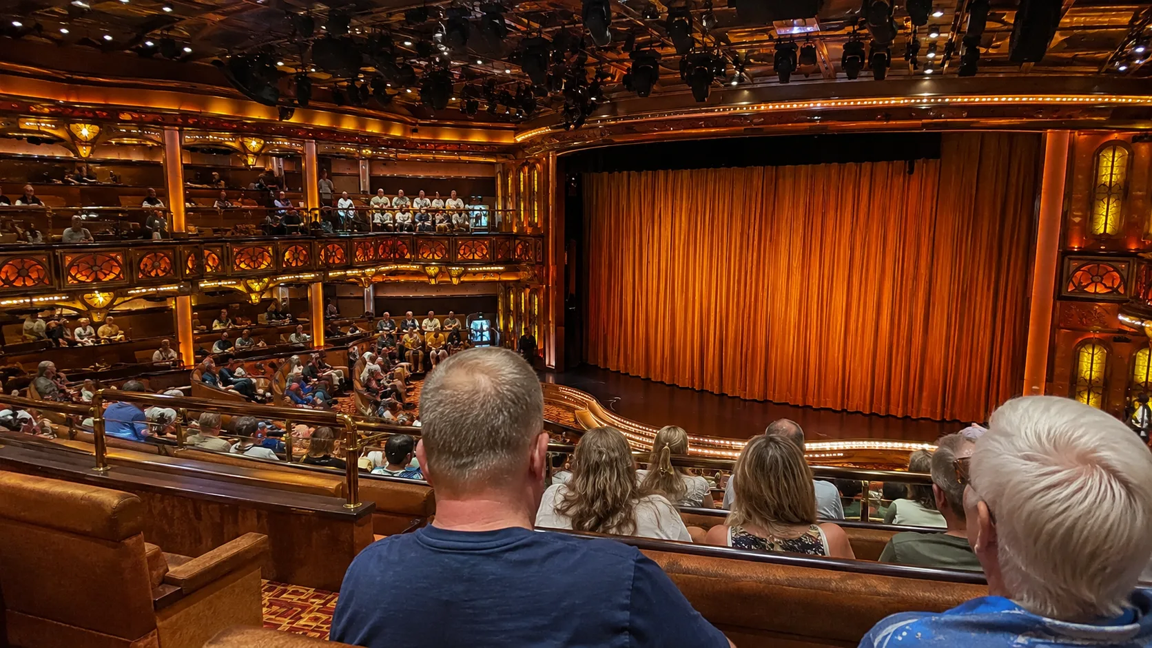 Audience seated before an orange stage curtain in an ornate cruise ship theater