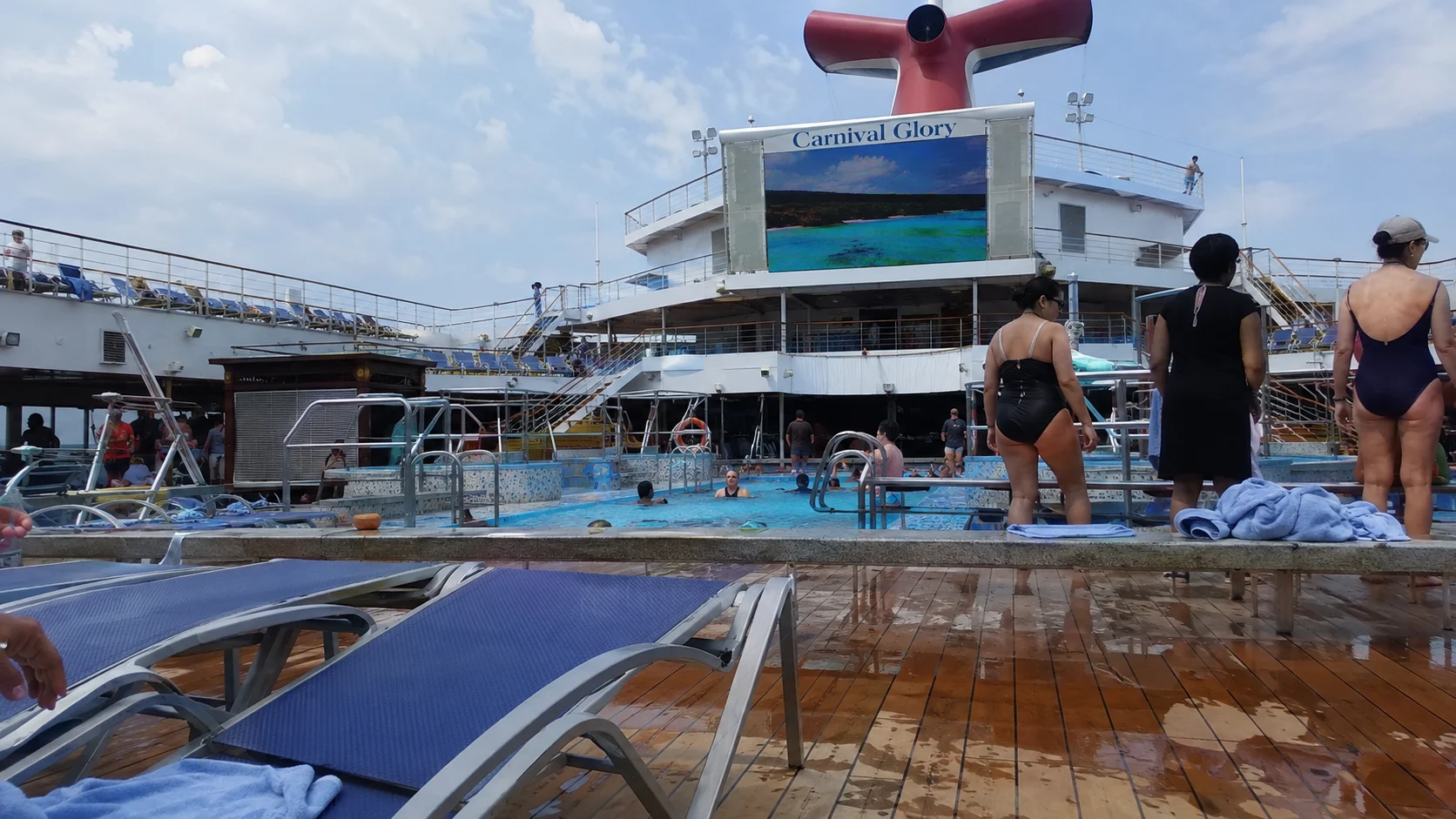Passengers beside a cruise ship pool beneath a large outdoor screen