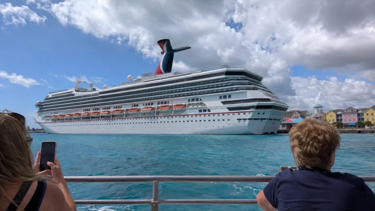 Passengers watching a Carnival cruise ship across turquoise harbor water