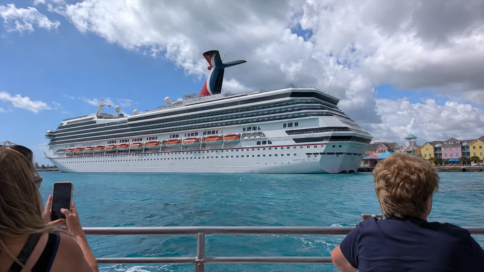Passengers watching a Carnival cruise ship across turquoise harbor water
