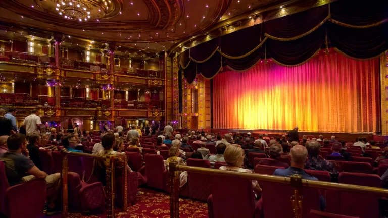 Audience seated before a red-and-gold stage curtain in an ornate theater