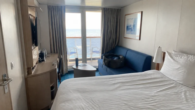 Cruise cabin with a double bed, blue sofa, and ocean-view balcony