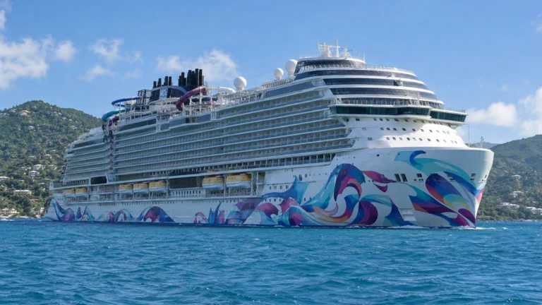 Norwegian Aqua sails past the green hills of St. Thomas, showing its colorful abstract hull art and upper-deck Slidecoaster.