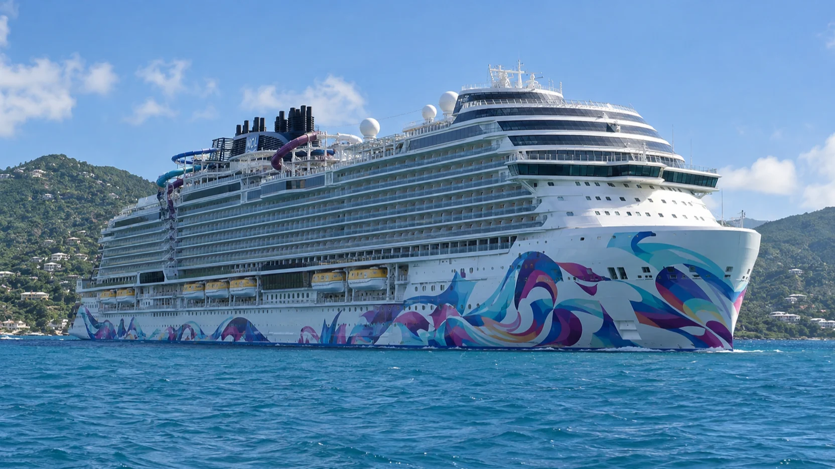 Norwegian Aqua sails past the green hills of St. Thomas, showing its colorful abstract hull art and upper-deck Slidecoaster.