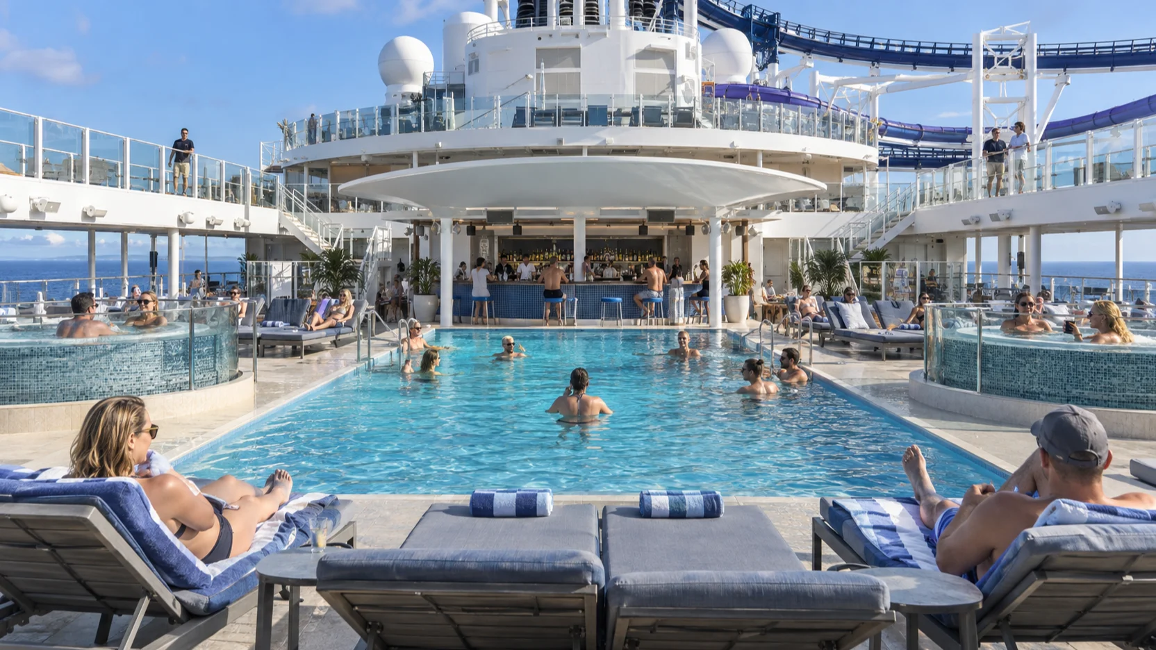Guests swim and lounge around Norwegian Aqua's main pool, raised hot tubs and Waves Pool Bar on a sunny sea day.